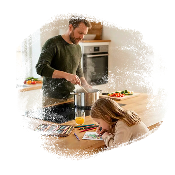 Pige sidder lænet ind over køkkenbord og farvelægger kort. Far står og røre i en gryde i baggrunden.